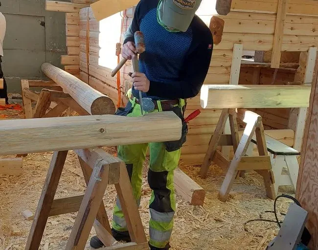 Elev arbeider med tømmerstokker på bukker ved et laftet hus